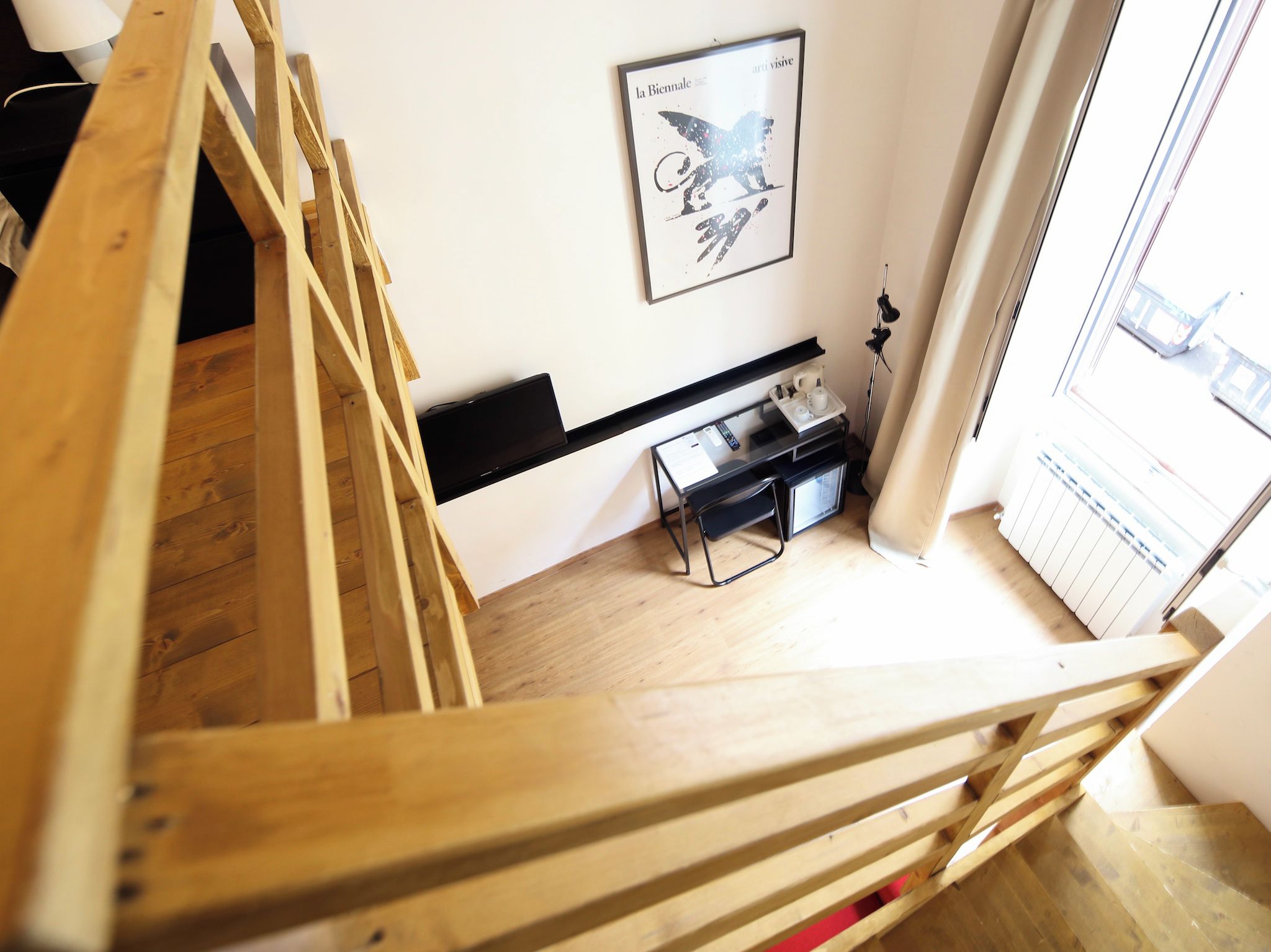 Un escalier en bois mène à une pièce où se trouve un bureau noir, un tableau encadré accroché au mur et une fenêtre donnant sur un paysage.