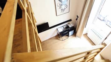 Un escalier en bois mène à une pièce où se trouve un bureau noir, un tableau encadré accroché au mur et une fenêtre donnant sur un paysage.