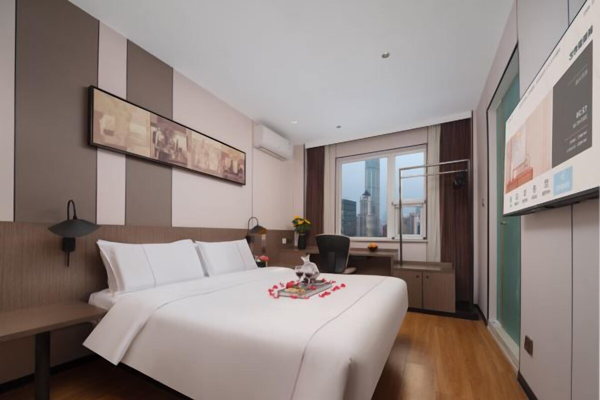 A hotel room with a bed, a desk, a chair, a window with a city view, and a wall-mounted artwork.