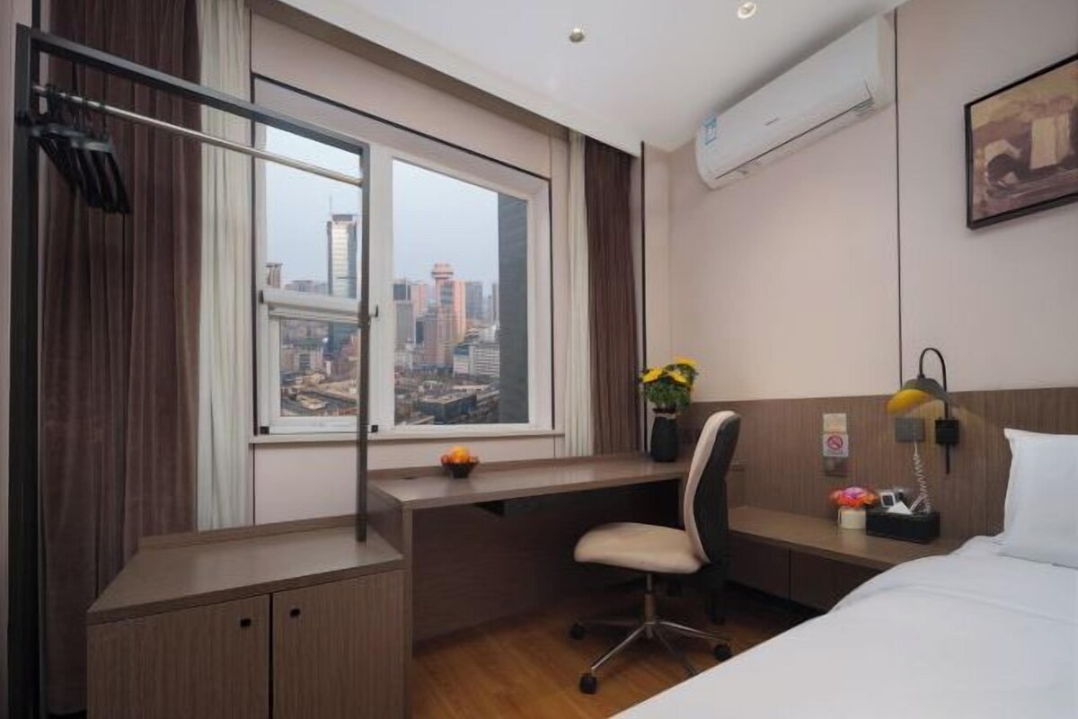 A hotel room with a desk, a chair, a bed, a window with a city view, and an air conditioning unit.