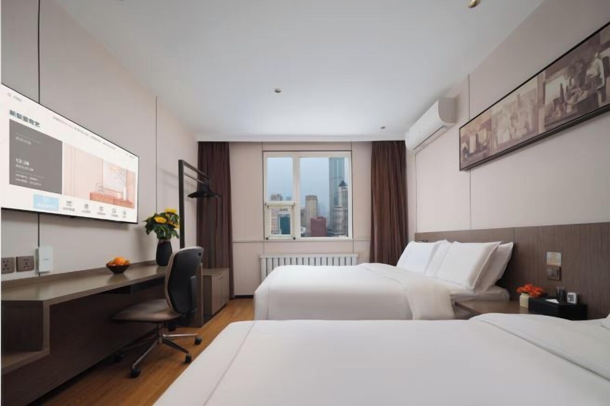A hotel room with a bed, a desk, a chair, a window with a view of buildings, and a wall-mounted screen.