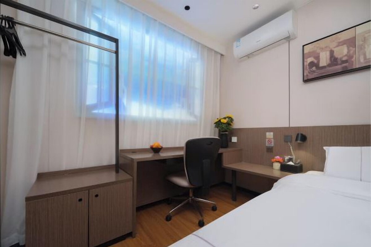 A hotel room with a bed, a desk, a chair, a window with sheer curtains, a wall-mounted air conditioner, and a framed picture.