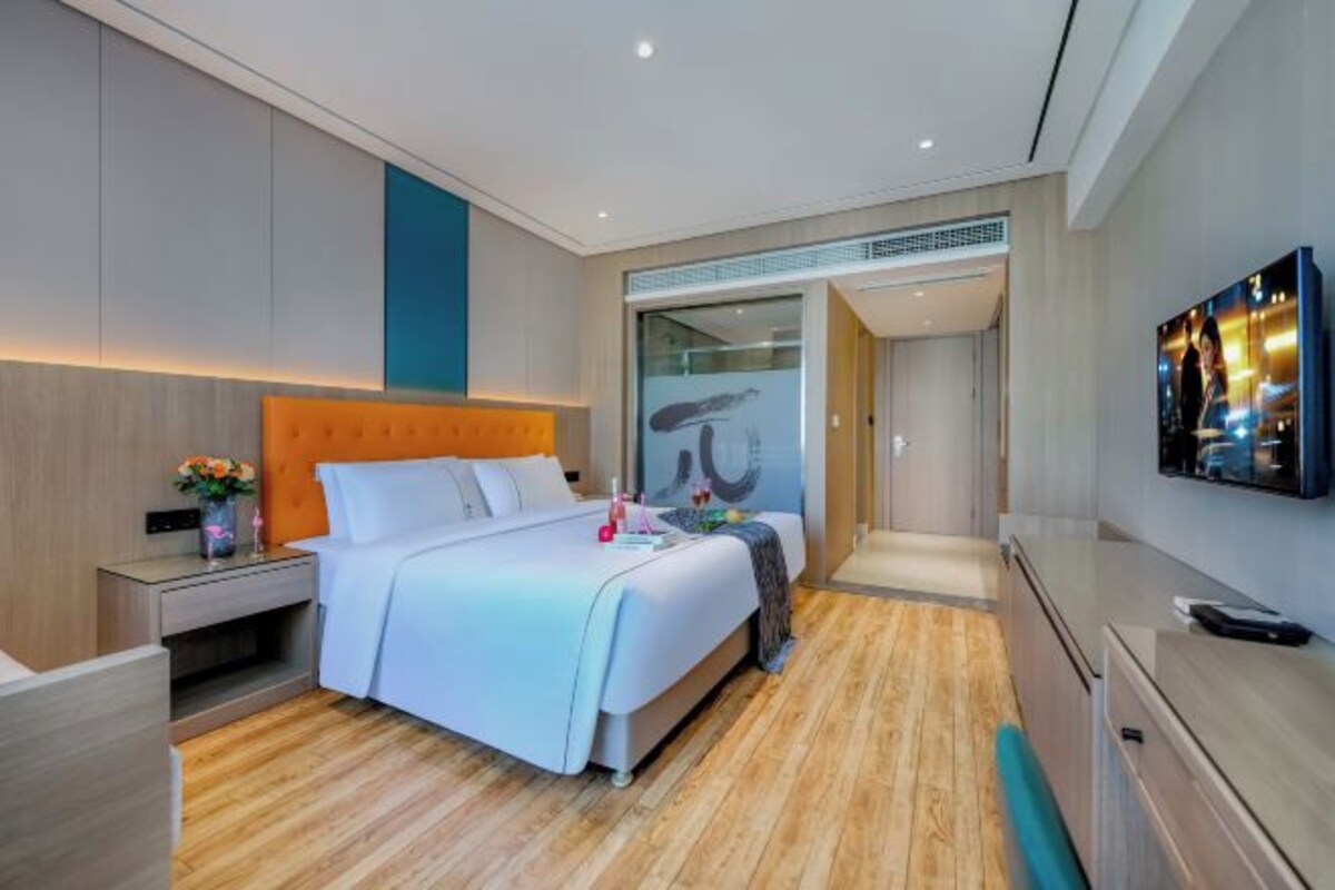 A modern hotel room with a large bed, a bedside table with a vase of flowers, a flat-screen TV mounted on the wall, and a wooden floor.