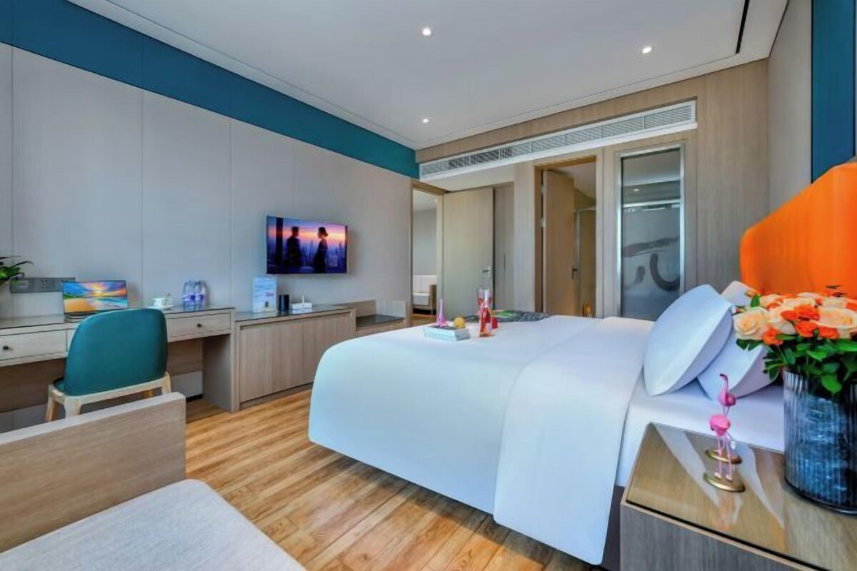 A modern hotel room with a large bed, a desk with a computer, a chair, and a TV.