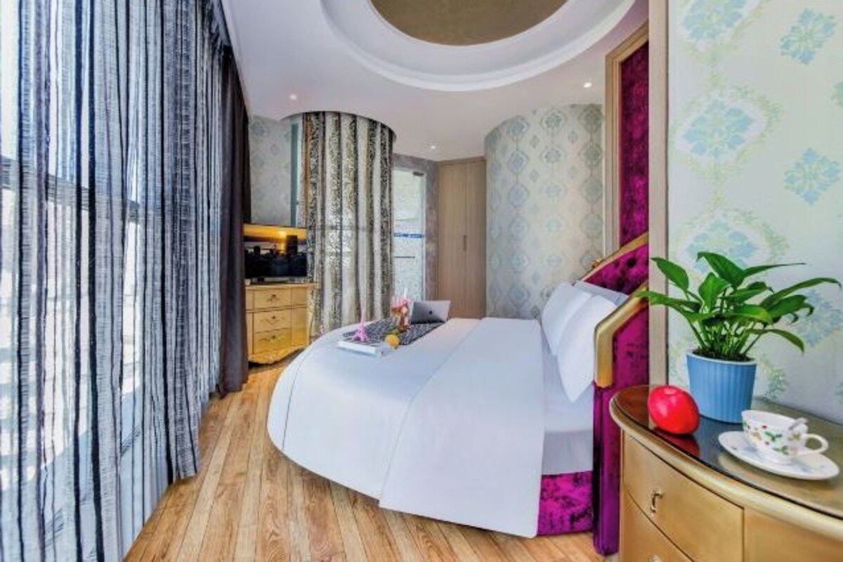 A modern bedroom with a curved bed, a wooden dresser, a potted plant, and a view of the city.