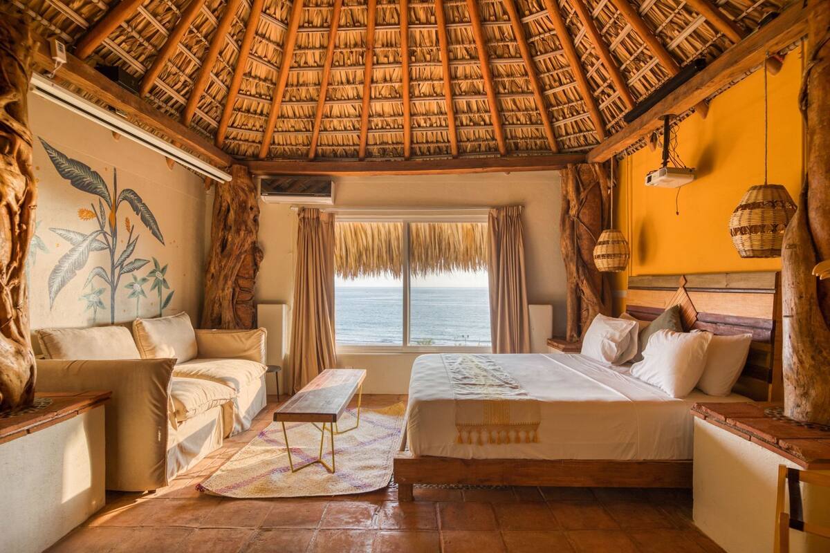 Una habitación con techo de paja, una cama, un sofá, una mesa y vistas al mar.