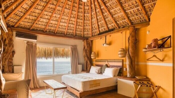 Ein Zimmer mit einem Reetdach, einem Bett, einem kleinen Tisch, einer Bank und Blick auf den Ozean.