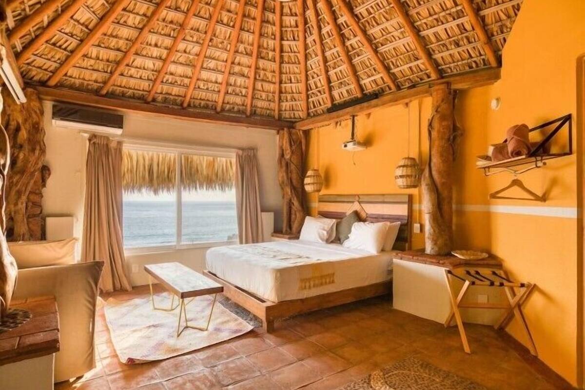Una habitación con techo de paja, una cama, una mesita, un banco y vista al océano.
