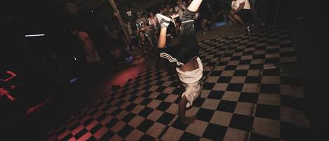 A person performing a handstand on a checkered floor in a dimly lit room with a crowd in the background.
