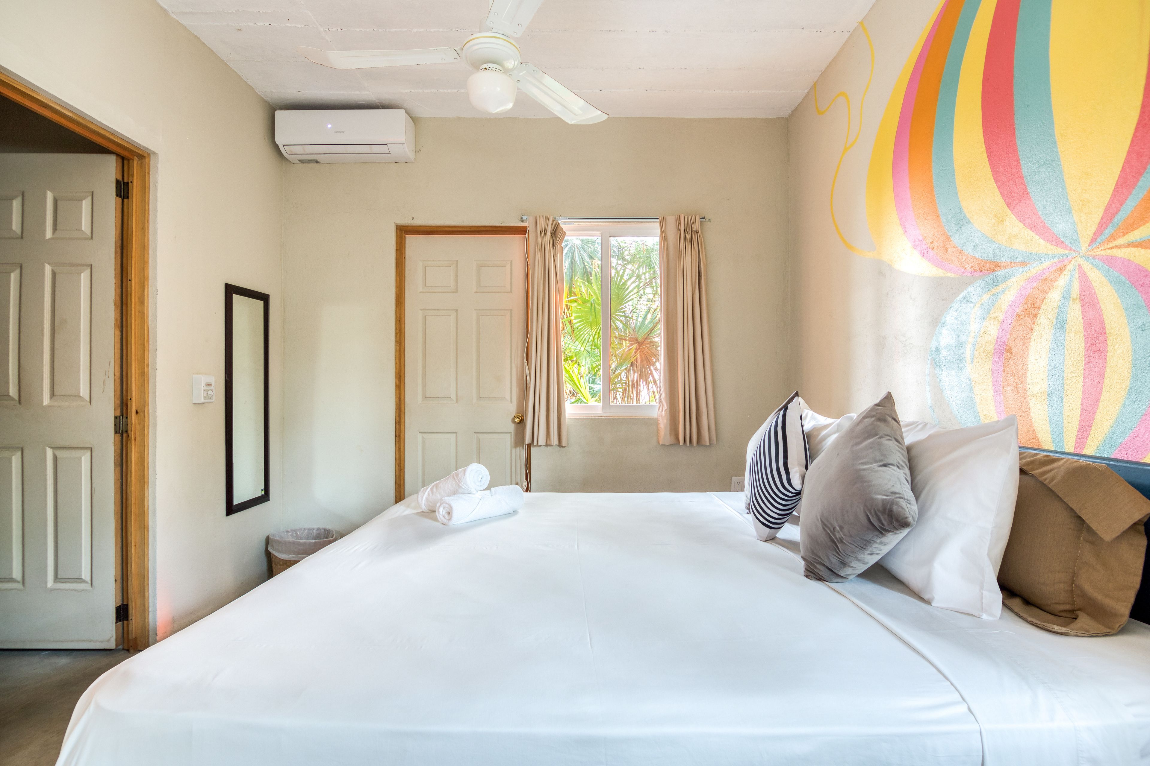 A hotel room with a large bed, a ceiling fan, a window with curtains, and a colorful wall mural.