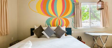 Habitación de hotel con cama, un mural de colores en la pared, una ventana con cortinas y una pequeña mesa con tabla de planchar.