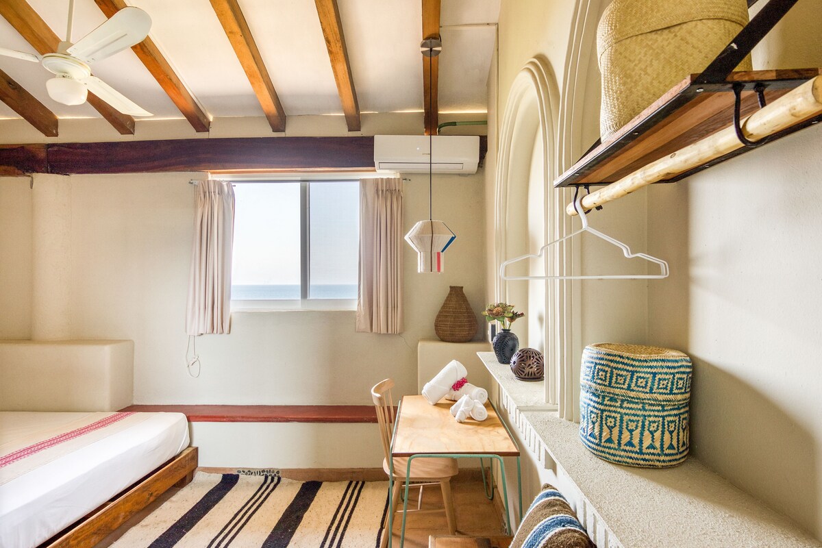 Une pièce comprenant un lit, une table, des chaises, un luminaire suspendu, une fenêtre avec des rideaux et une étagère ornée d’objets décoratifs.