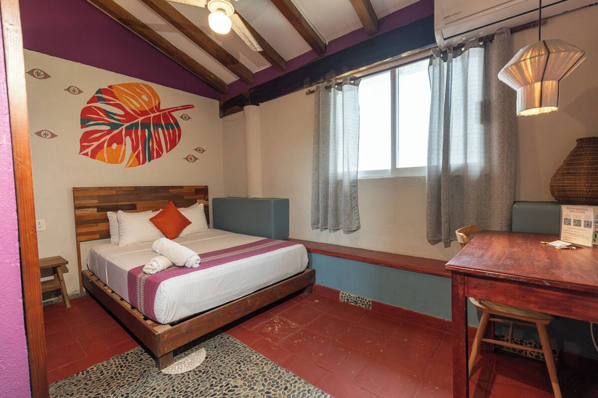 Un dormitorio con cama, cabecero de madera, ventana con cortinas, una mesa y pared con diseño de hojas.