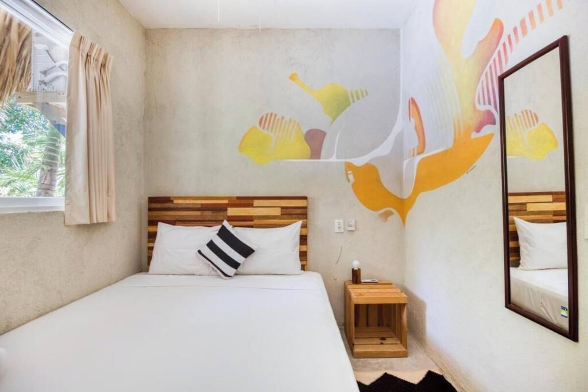 Un dormitorio con cabecera de madera, cama con ropa de cama blanca, mesita de noche, espejo montado en la pared y un mural.