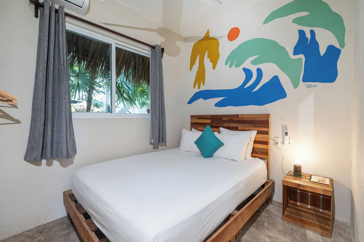 Una cama con cabecera de madera, una mesita de noche con lámpara y un mural en la pared.