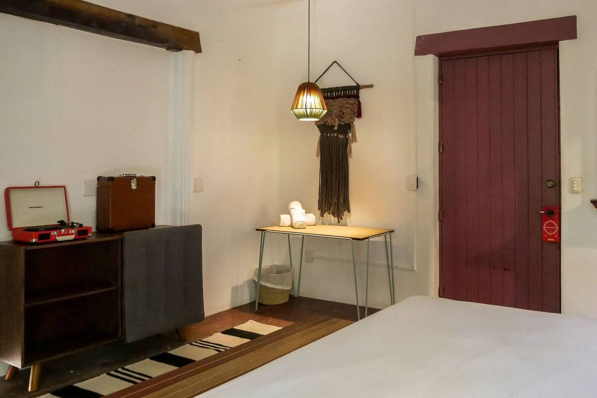 Una habitación con una cama, un escritorio con lámpara, una maleta y una puerta roja.