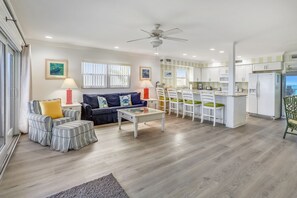 Un salon moderne comprenant un canapé, un pouf et une table basse, un coin repas avec des chaises, et une cuisine équipée d’un réfrigérateur et d’un four à micro-ondes.