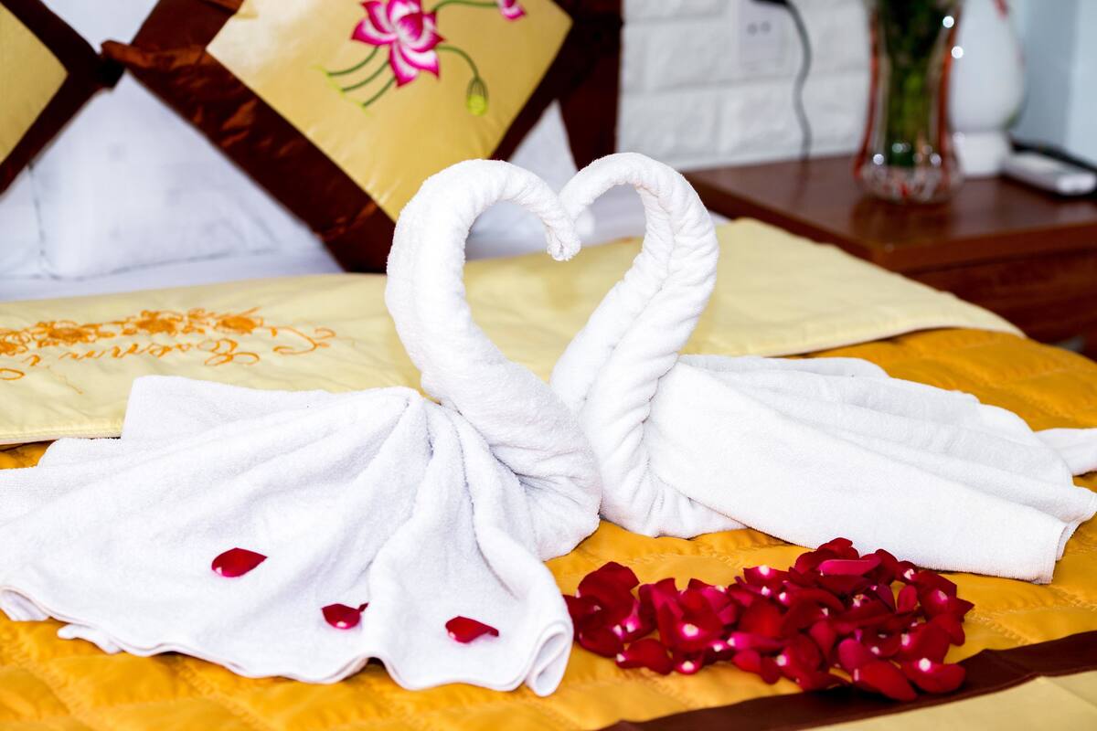 A bed with a yellow bedspread, two white towels arranged in a heart shape, and scattered red rose petals.
