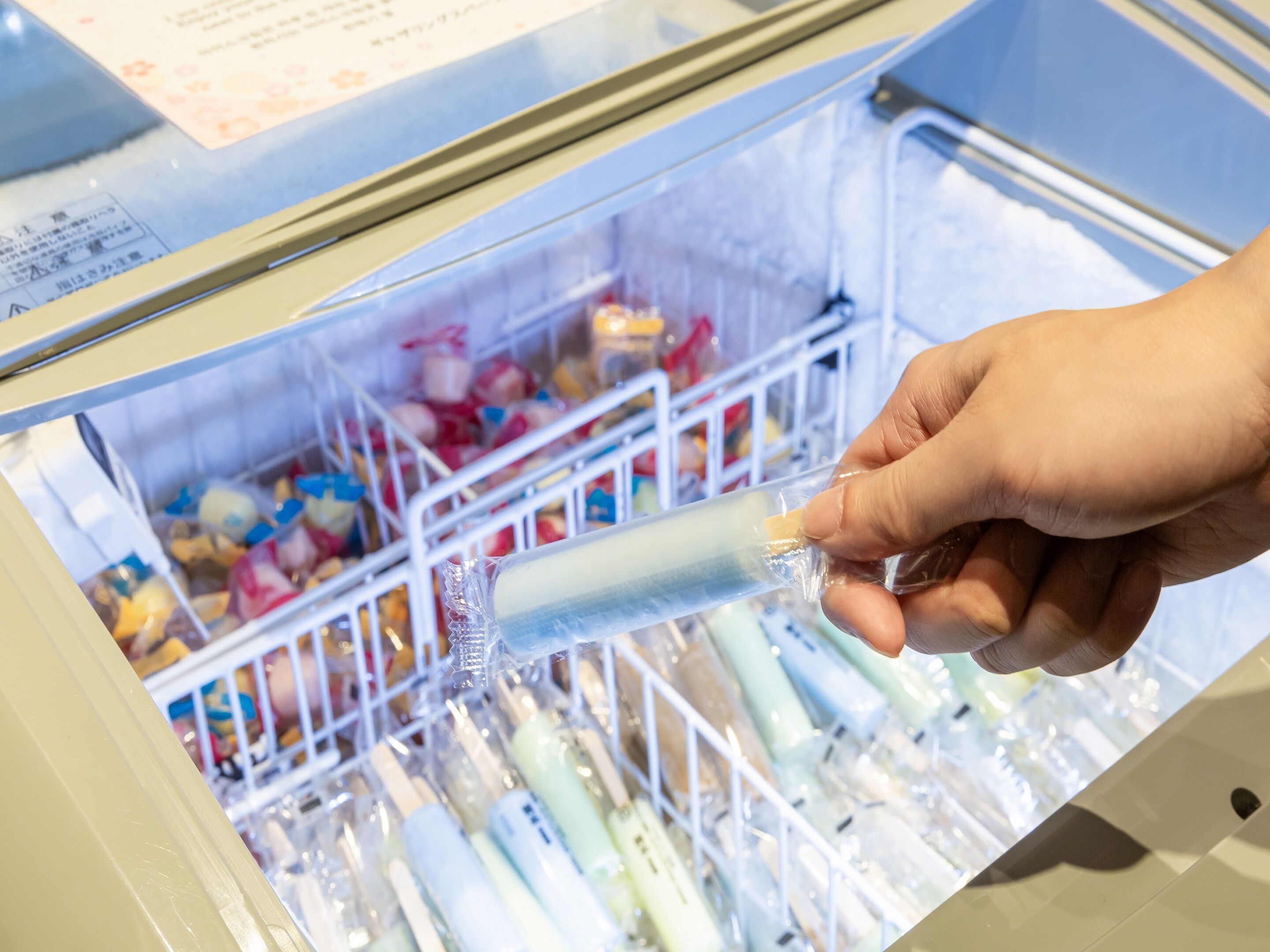 A hand holding a sealed tube in a refrigerated display case with multiple tubes and colorful items in the background.