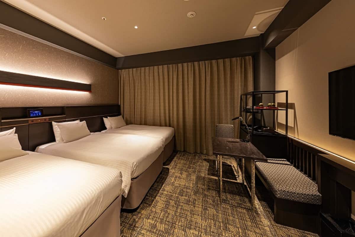 Ein Hotelzimmer mit zwei Betten, einem Schreibtisch, einem Stuhl und einem an der Wand befestigten Fernseher.