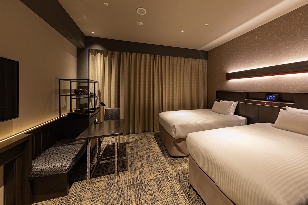 Ein Hotelzimmer mit zwei Betten, einem Schreibtisch mit Stuhl, einem Fernseher und einer Bedienkonsole an der Wand.