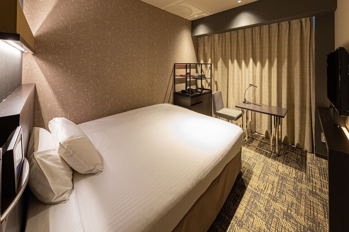 Ein Hotelzimmer mit einem großen Bett, einem Schreibtisch mit Stuhl, einem Fernseher und einem Fenster mit Vorhängen.
