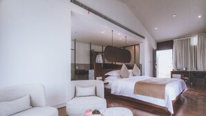 Una habitación de hotel moderna con una cama grande, un sofá, una mesa de comedor con una bandeja de fruta y té, y vistas al exterior.