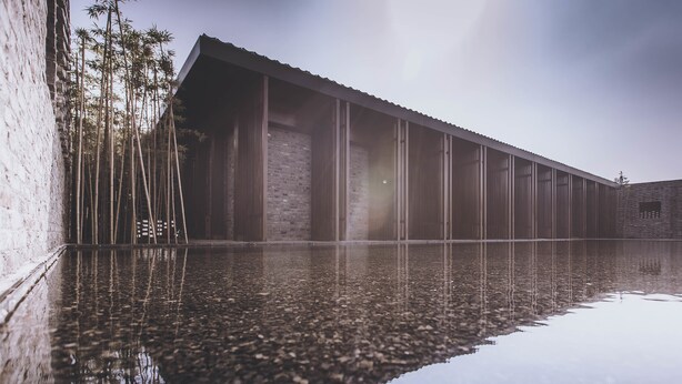 Un edificio moderno con una piscina reflectante, árboles de bambú y un muro de ladrillo.