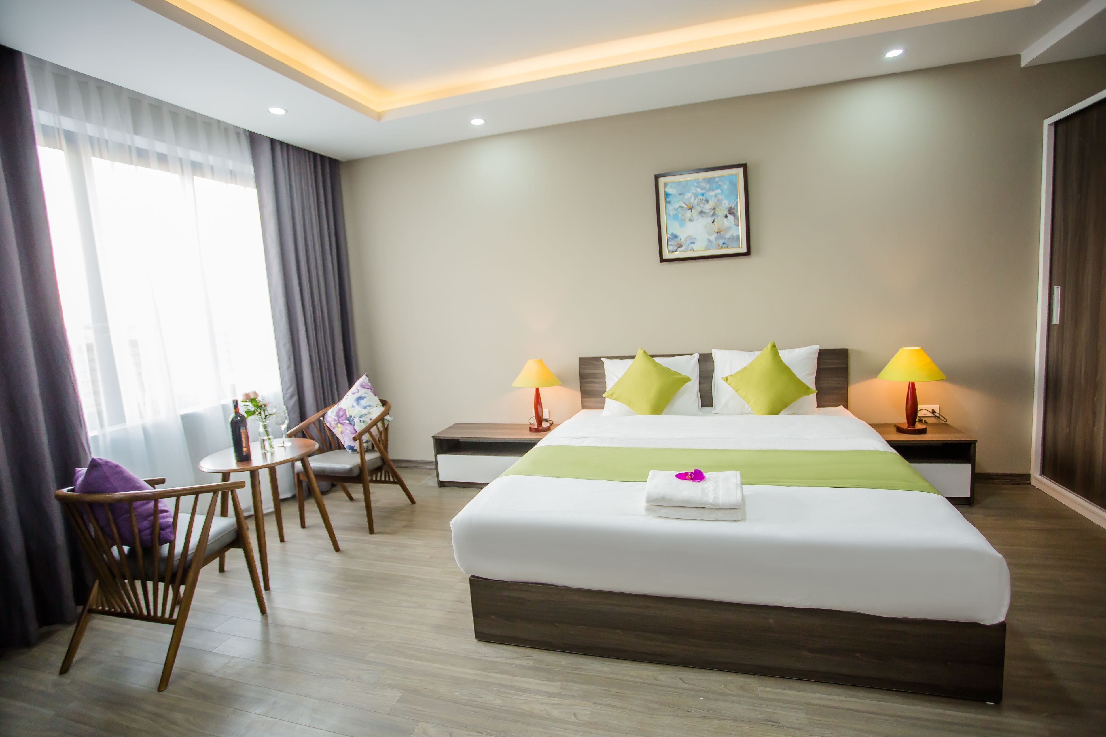 Una habitación de hotel moderna con una cama grande, dos mesitas de noche con lámparas, una pequeña mesa con una silla y un amplio ventanal con cortinas.