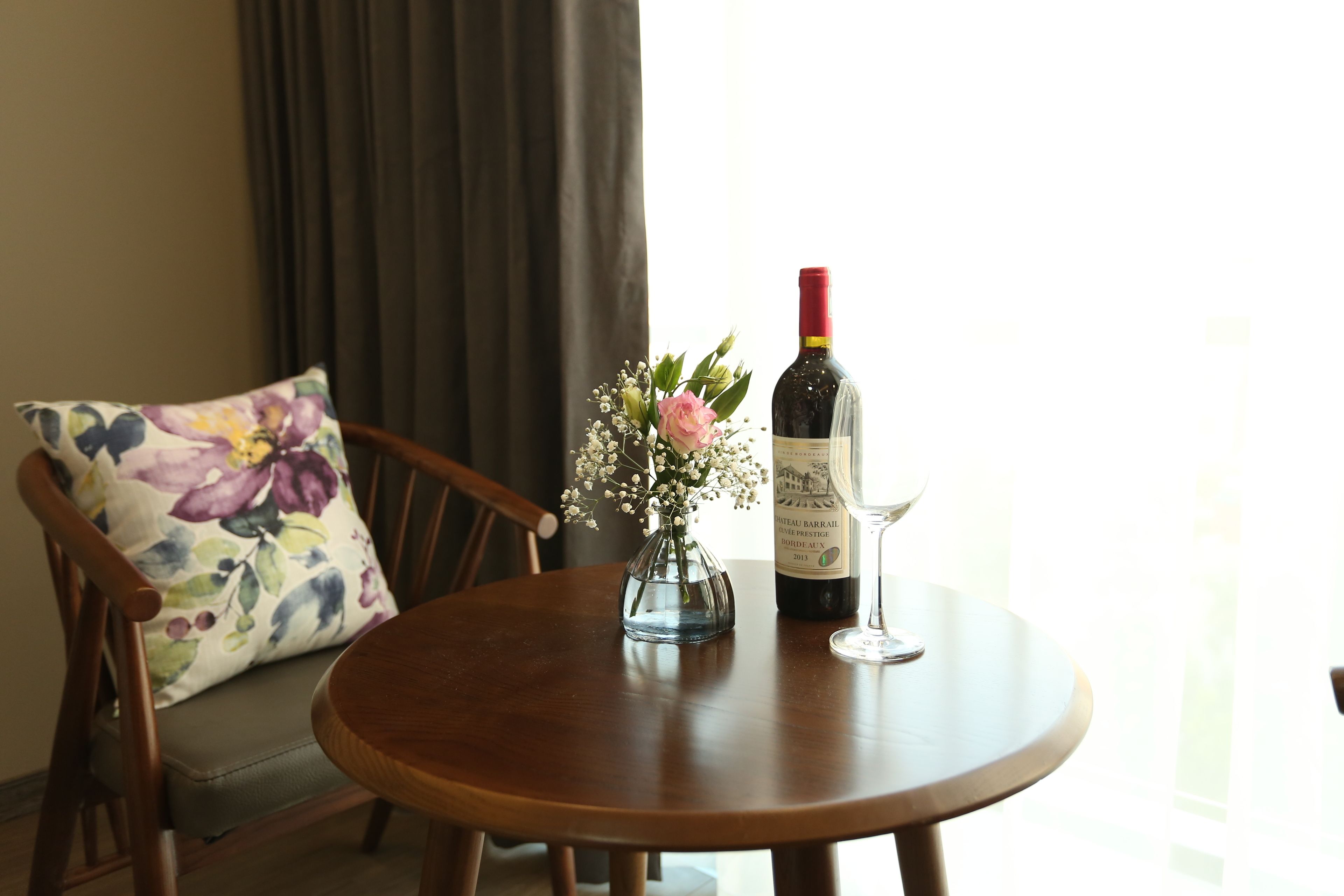 Een ronde houten tafel met een wijnfles en glas, een bloemenkussen en een vaas met bloemen.