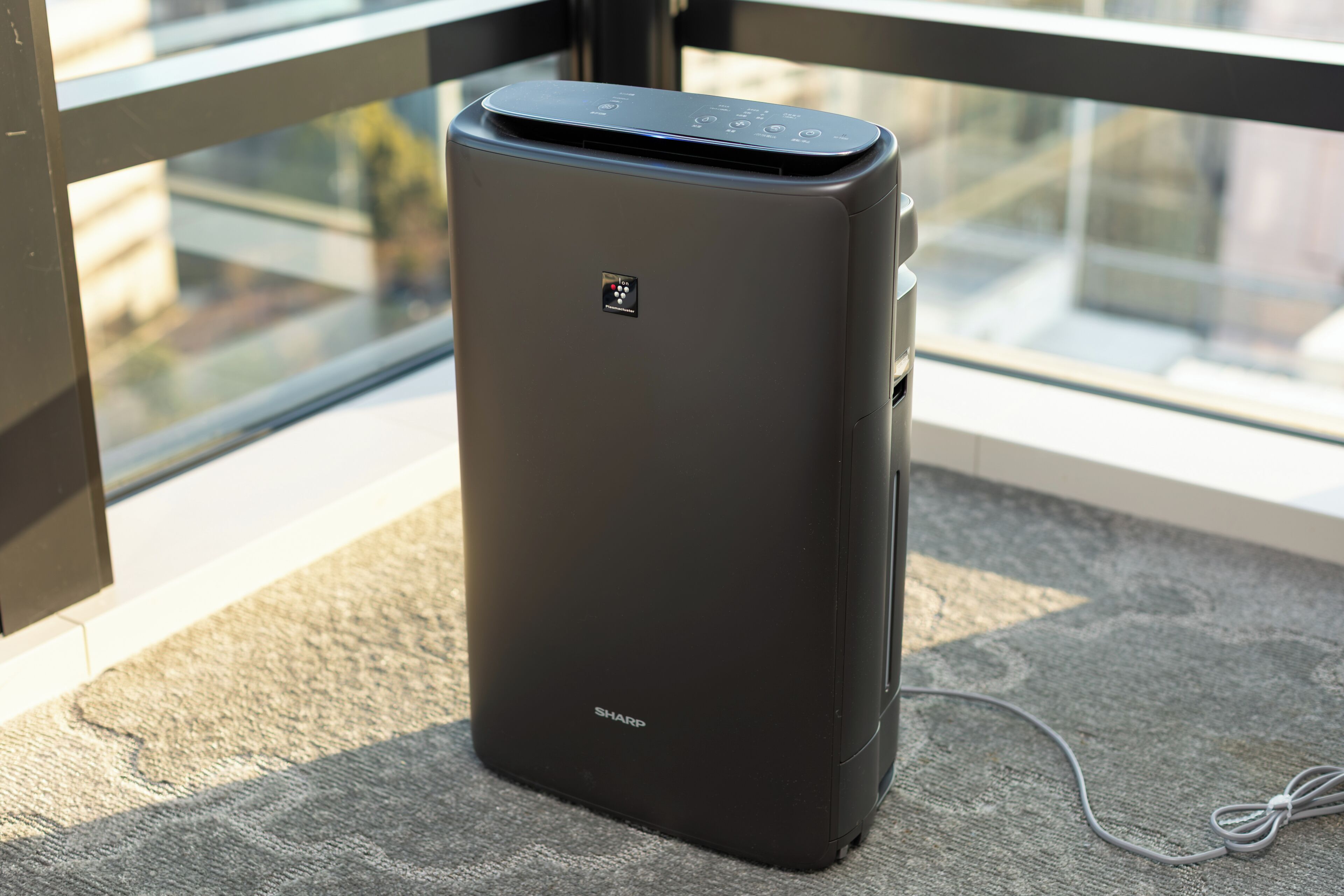 A black Sharp air purifier on a carpeted floor near a window.