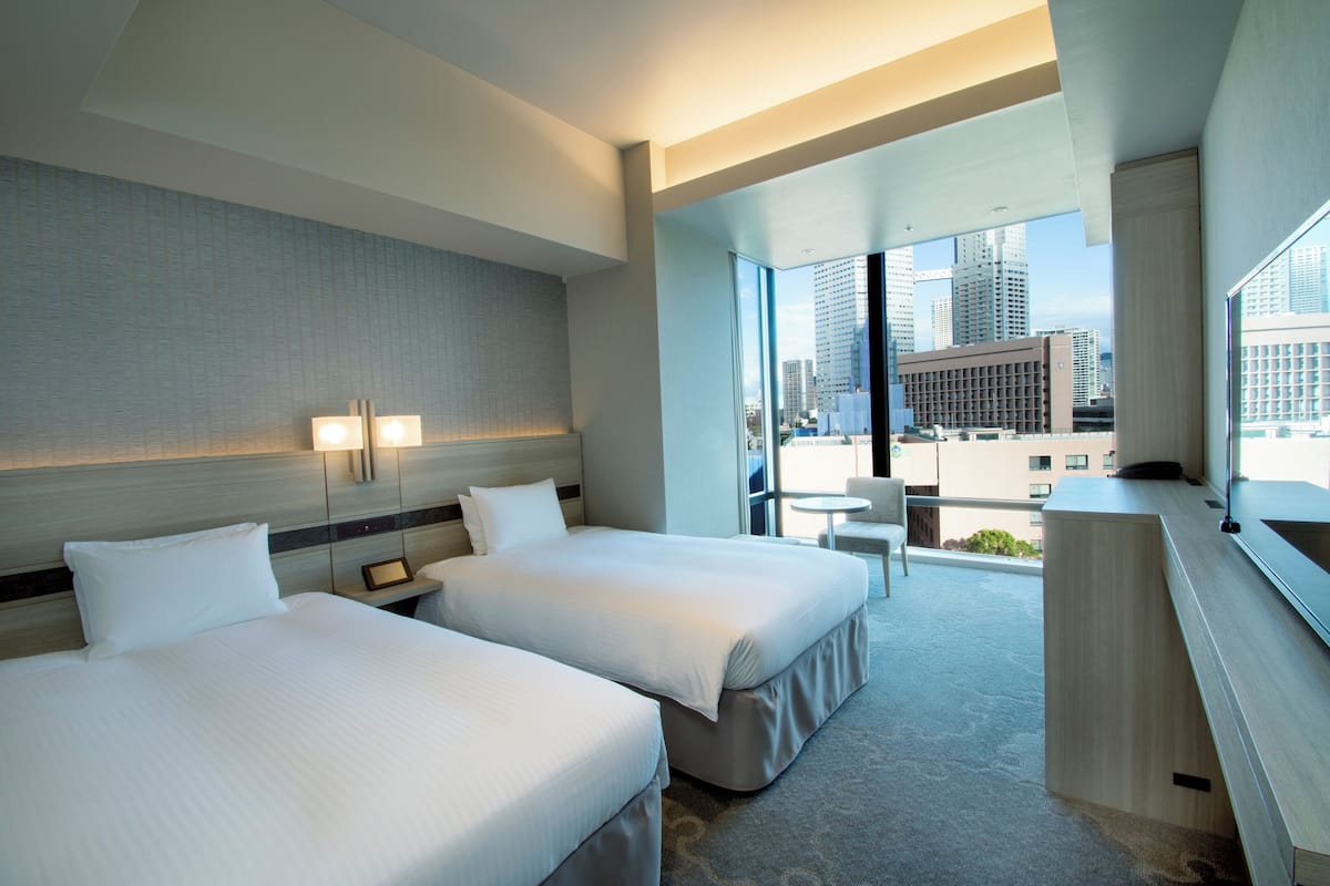 A hotel room with two beds, a desk, a chair, and a large window offering a city view.
