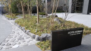 A landscaped area with a sign for Hotel Keihan, featuring a modern building in the background. - Hotel Keihan Tsukiji Ginza Grande (Tokyo)