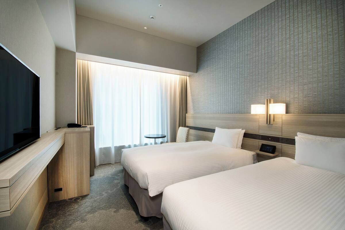 A hotel room with two beds, a TV, a desk, and a window with curtains.