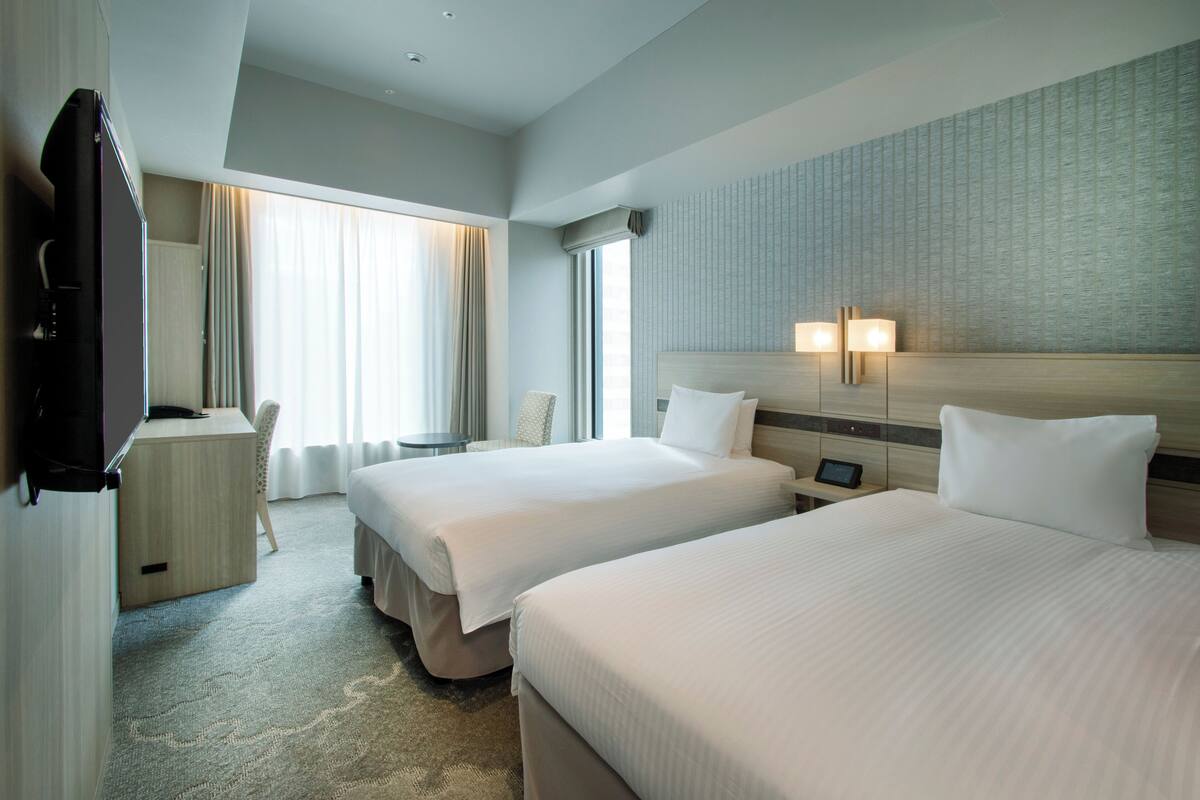 A hotel room with two beds, a desk, a chair, and a TV.