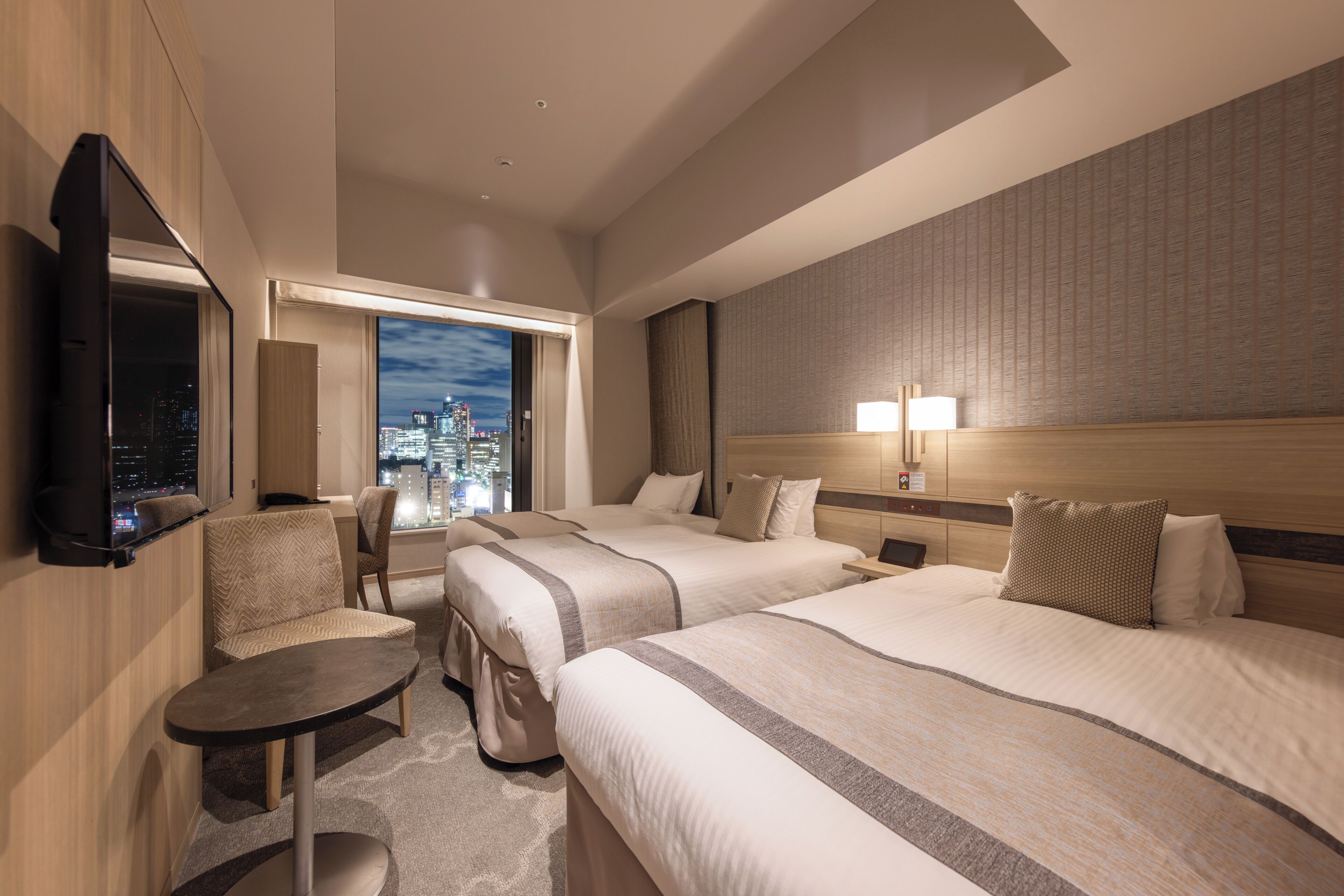 A hotel room with two beds, a TV, a small table, and a view of a cityscape at night.