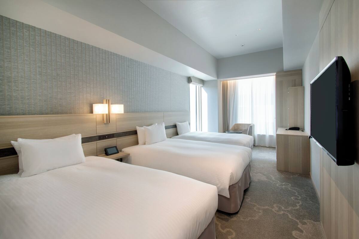 A hotel room with three beds, a flat-screen TV, and a large window with curtains.