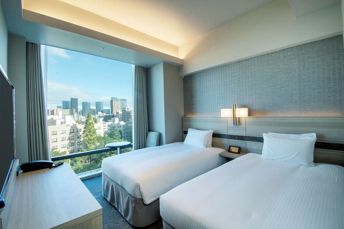 A hotel room with two beds, a desk, a chair, and a large window offering a city view.