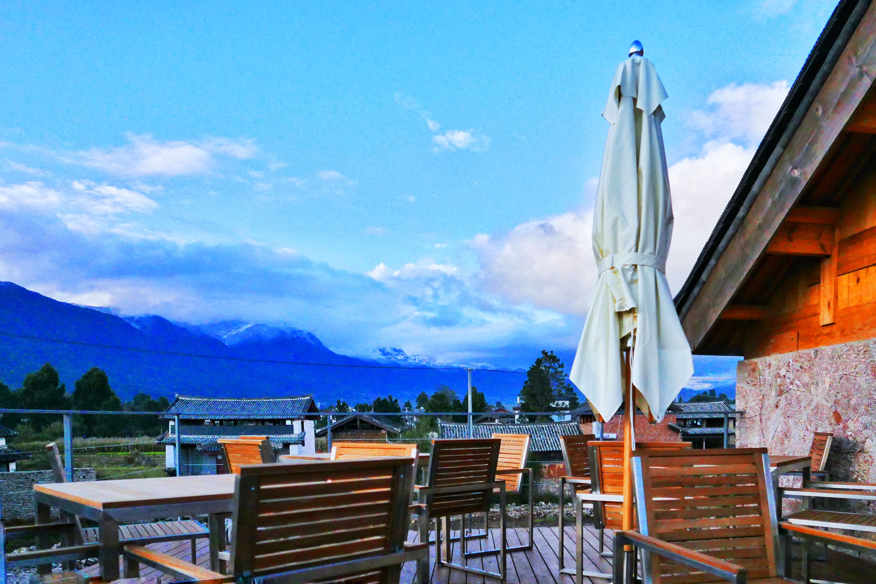 An outdoor seating area with wooden tables and chairs, a white umbrella, and a view of a mountain range.