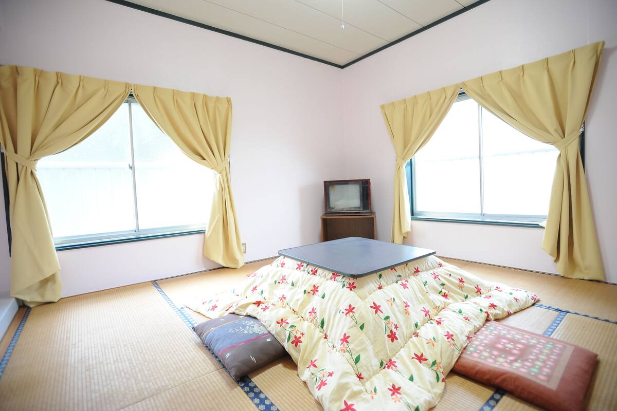 A room with tatami mat flooring, a low table, and two windows with curtains.