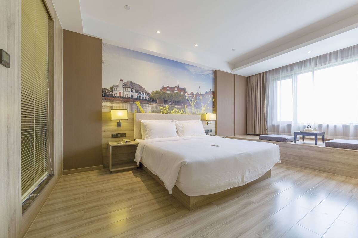 Ein Hotelzimmer mit einem großen Bett, einem Nachttisch, einer Lampe, einem Stadtgemälde und einem großen Fenster mit Vorhängen.