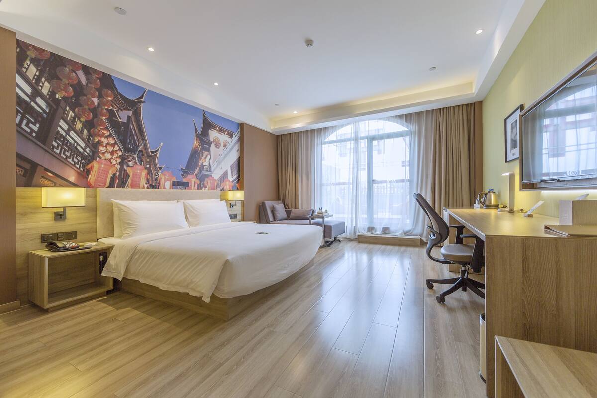 Ein Hotelzimmer mit einem großen Bett, einem Schreibtisch, einem Stuhl, einem Fernseher und einem großen Fenster mit Vorhängen.