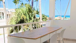 Un balcón con mesa de madera y sillas blancas, con vista a una playa con palmeras y un cielo azul despejado.