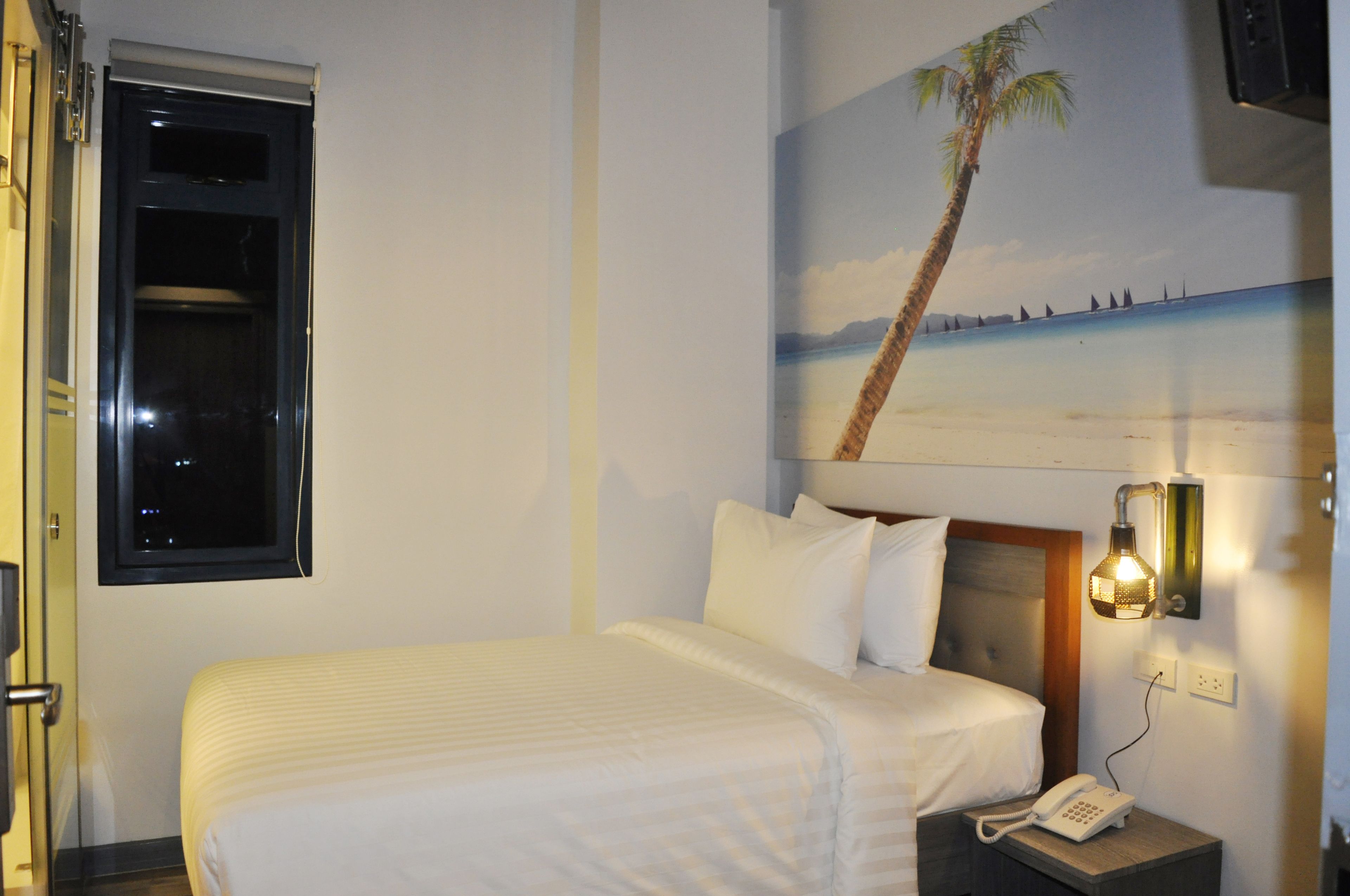 A hotel room with a bed, a nightstand with a phone, a wall-mounted light, and a window with a view of the beach.