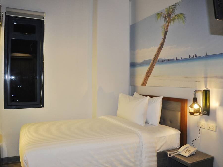 A hotel room with a bed, a nightstand with a phone, a wall-mounted light, and a window with a view of the beach.