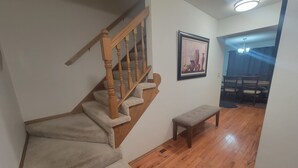 A staircase with carpeted steps and wooden railings, a framed picture on the wall, a bench, and a dining area with chairs and a table.