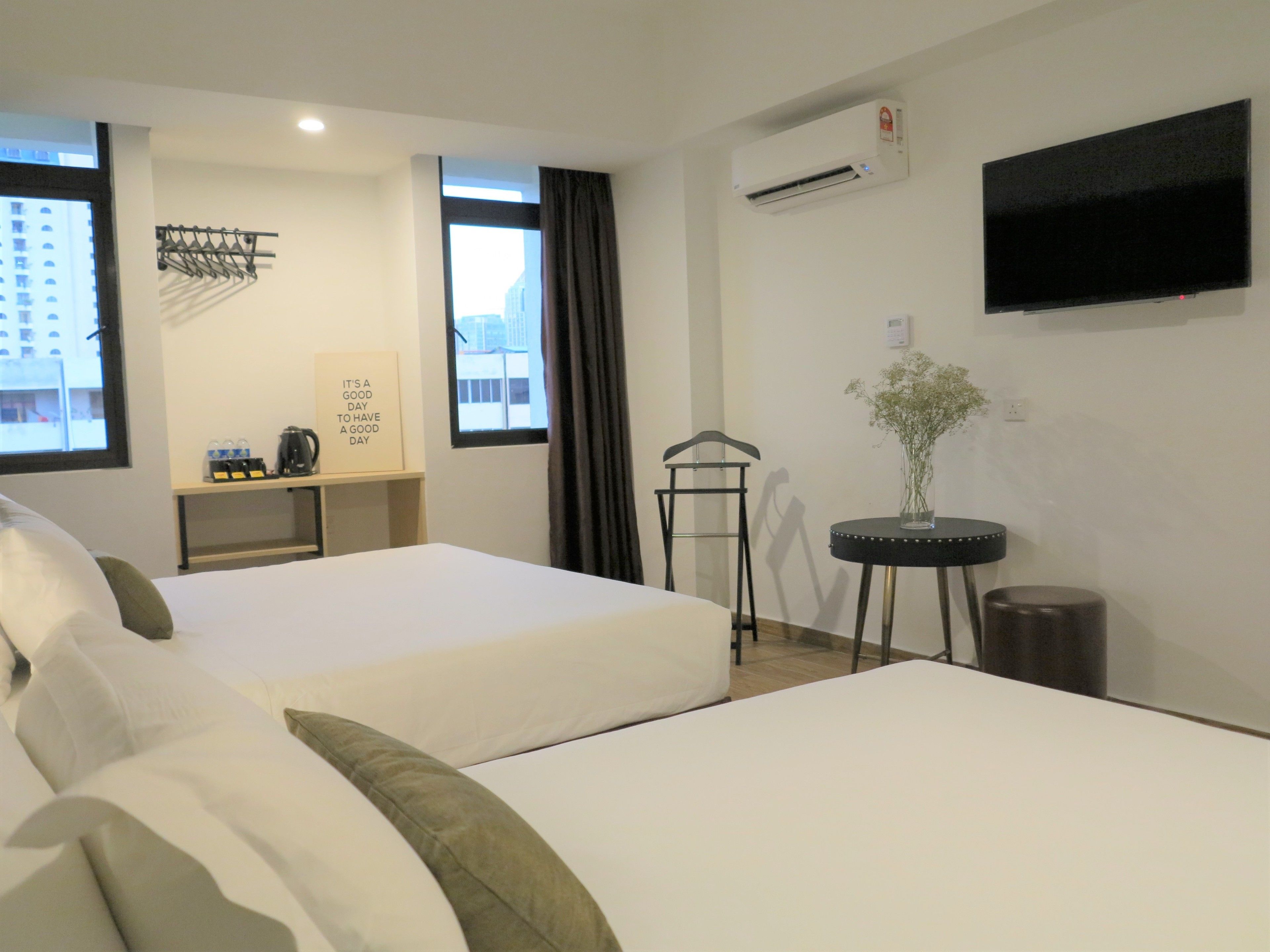 A hotel room with two beds, a TV, an air conditioner, a small table with a vase, and a window with a city view.
