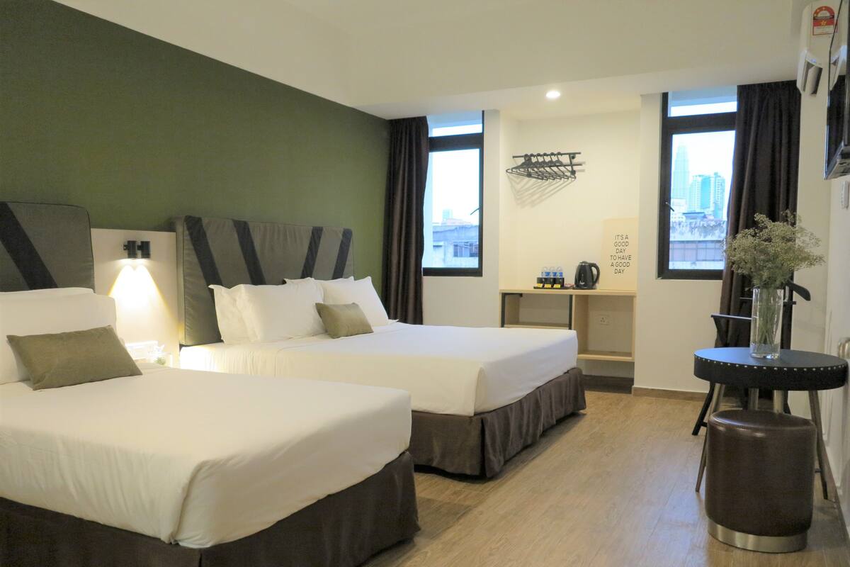 A hotel room with two beds, a small table with a kettle, a chair, and a view of the city through the windows.