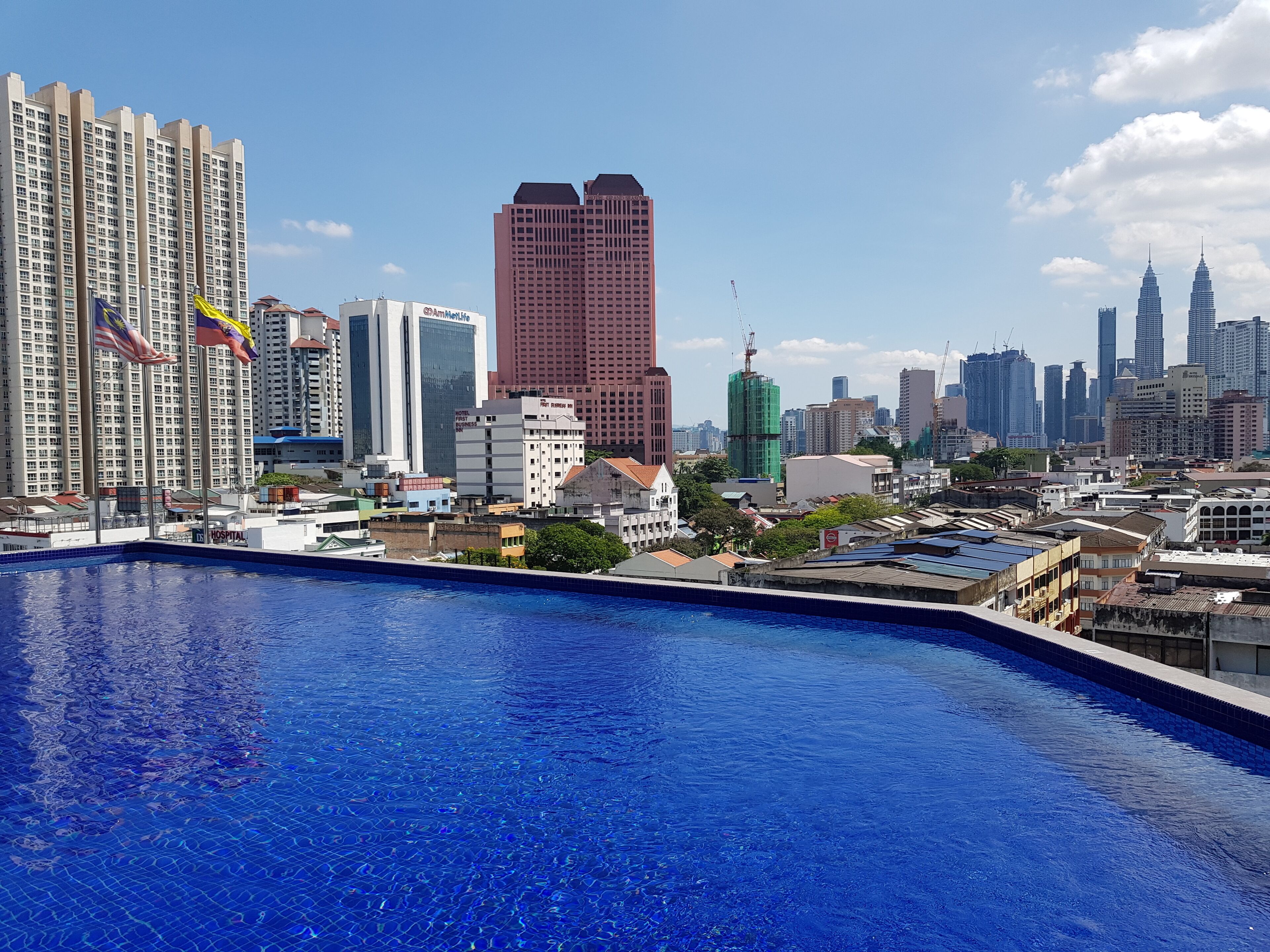 Un paisaje urbano con una gran piscina en primer plano, rodeado de rascacielos, incluyendo un hotel destacado de diseño moderno.