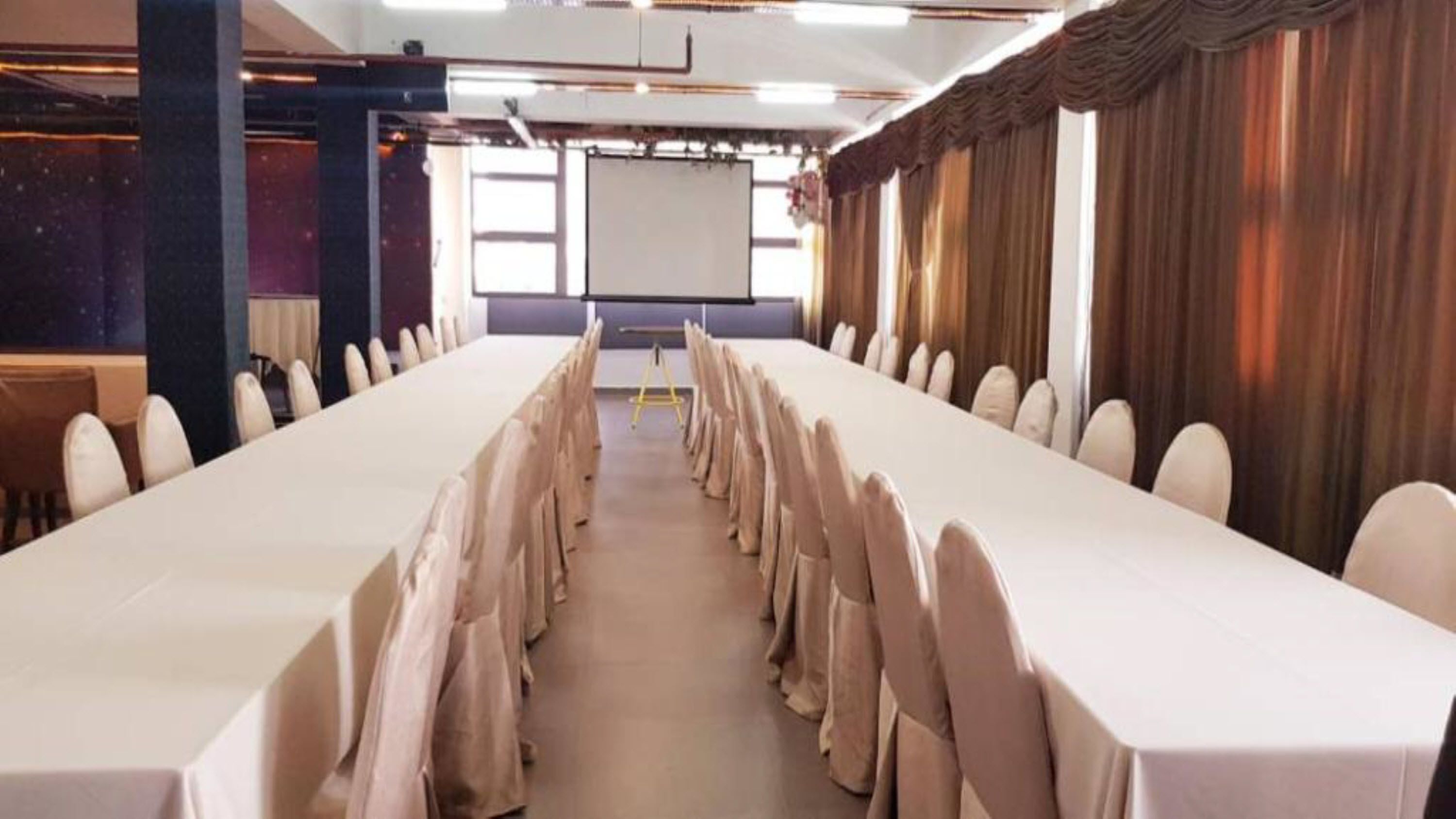 Un salón de conferencias con una larga mesa, filas de sillas, una pantalla de proyector y cortinas.